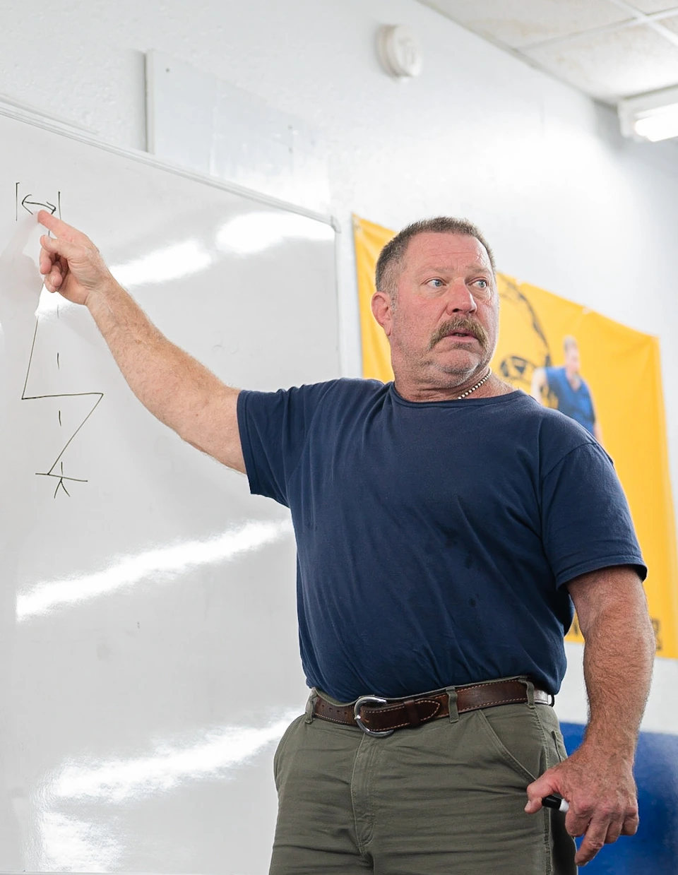 Mark Rippetoe teaching in a gym classroom, pointing at a whiteboard.