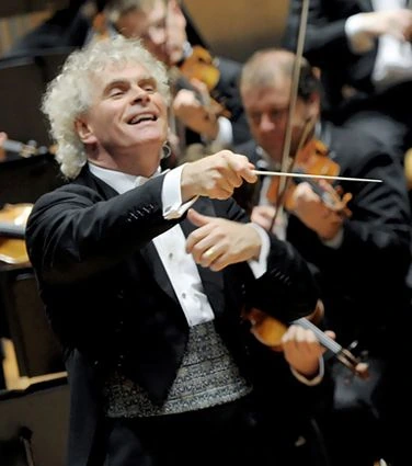 Sir Simon Rattle conducting an orchestra on stage.