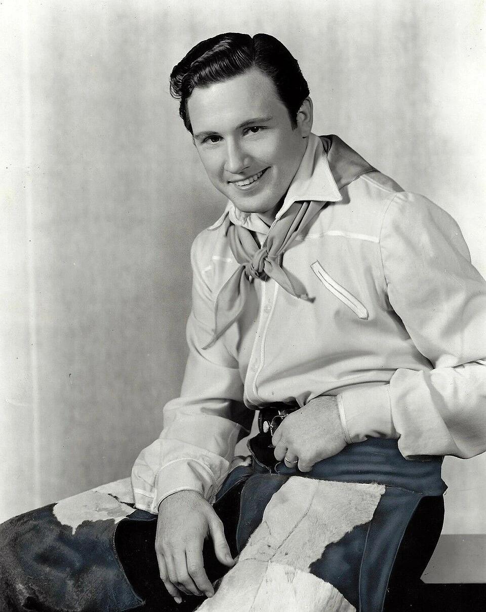 Black-and-white publicity portrait of John Raitt in a Western costume, smiling at the camera.